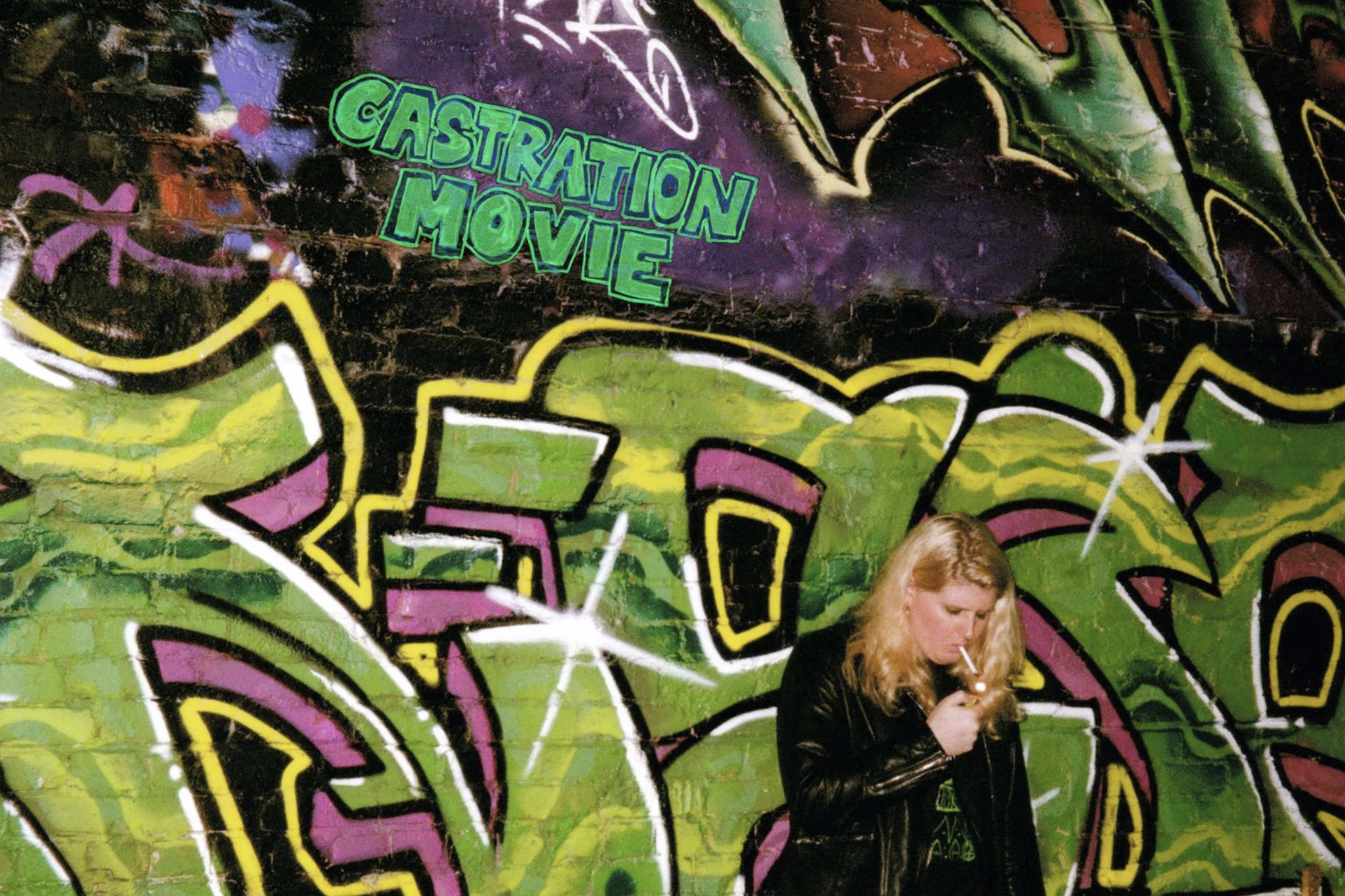 Louise Weard is smoking in front of a wall with bright green and purple graffiti. Castration Movie is written in green on top of the graffiti.