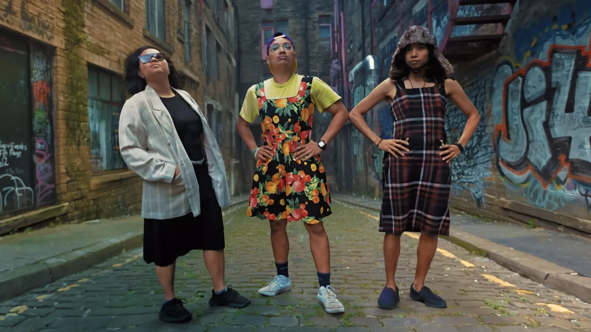 Three queer people stand in the middle of a street wearing colourful stylish clothes.