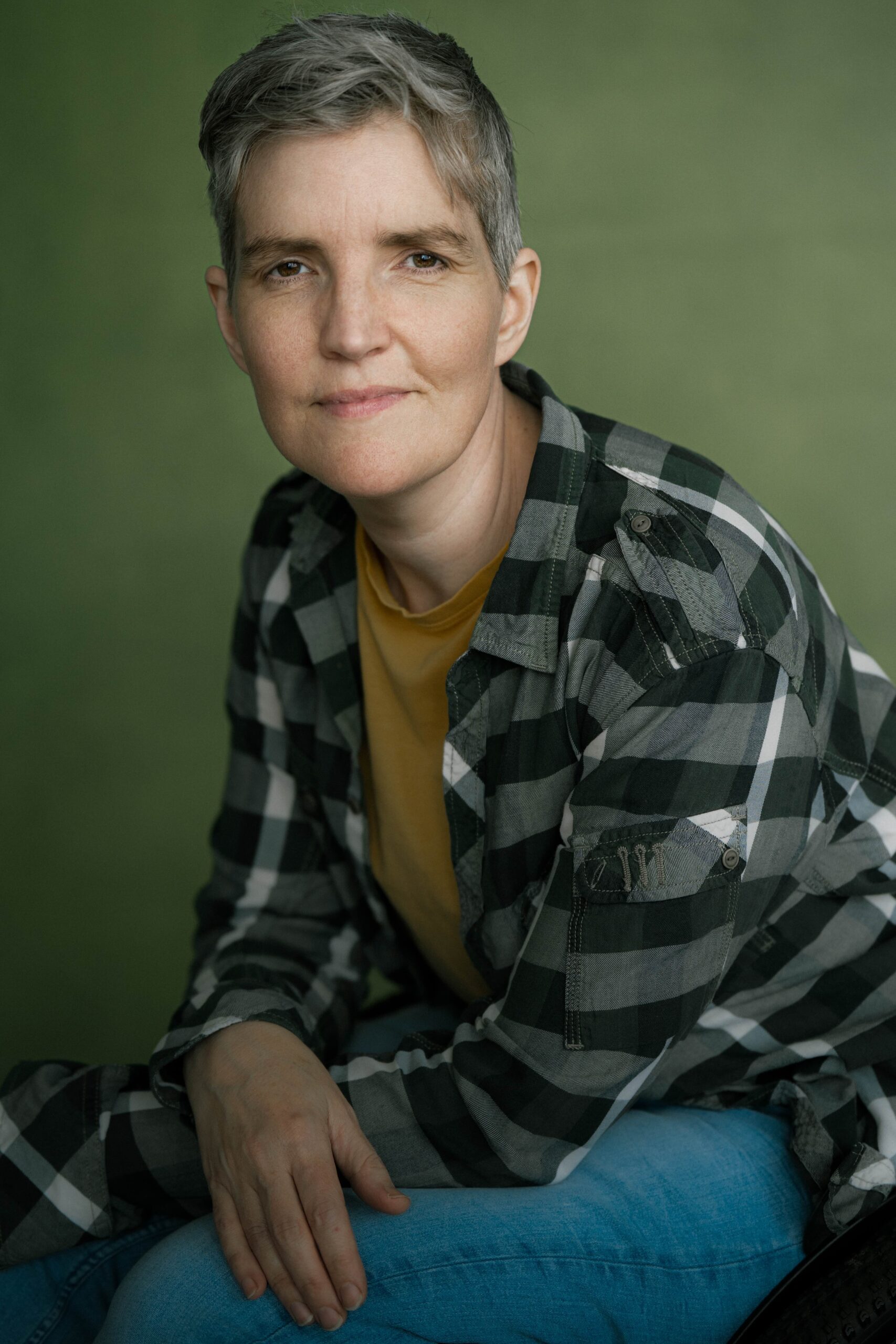 A closeup headshot of a non-binary white person in their early 40s, with short grey hair in various hues. They are looking straight ahead to the camera with a mainly neutral expression. They are wearing a green and white plaid shirt and posing against a red background.