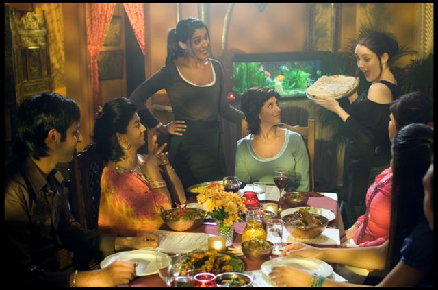 A South Asian family of 5 people enjoys dinner together with a white lesbian carrying a dish.