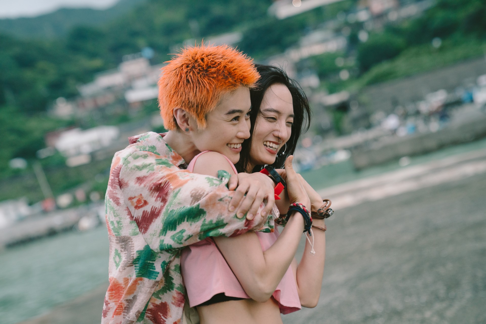 Two people smile and laugh as one embraces the other from behind on a beach. One has short, bright orange hair and wears a patterned shirt; the other wears a pink top. They stand close to the shoreline, with green hills and buildings blurred in the background.