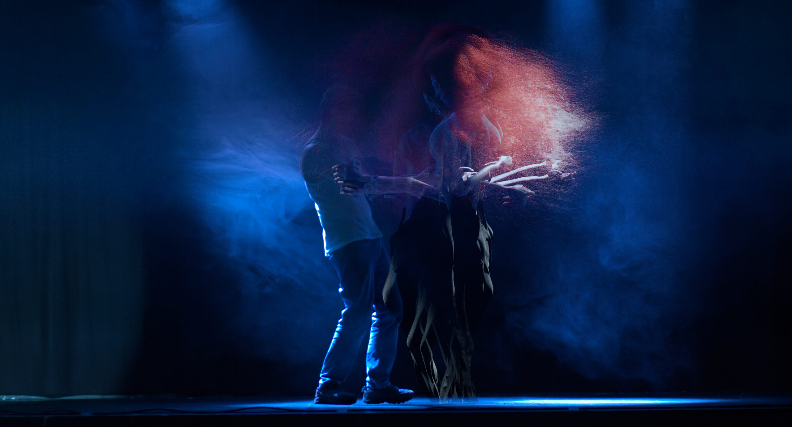 Two figures stand embracing on a dark stage, lit by blue spotlights and surrounded by smoke. One figure is a white man wearing a vest and jeans. The other is tall, semi-naked, with long ginger hair, digitally animated and superimposed several times in slightly different poses.