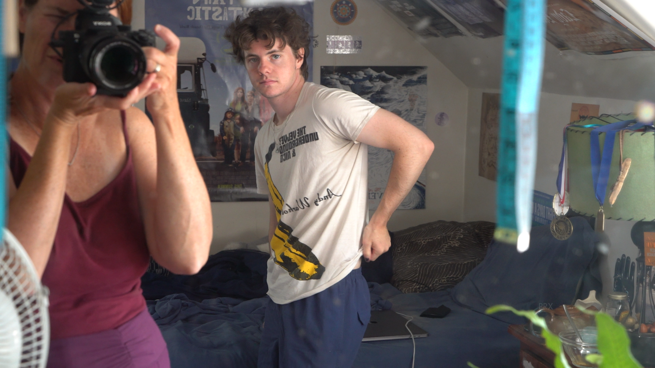 A young man with brown hair stands in a bedroom, caught in a candid pose, as he adjusts the hem of his t-shirt. In the foreground, a woman with reddish hair holds up a camera photographing him through a mirror. The room behind him is filled with personal touches: film posters on the wall, a rumpled navy bedspread, and medals hanging near the doorway.