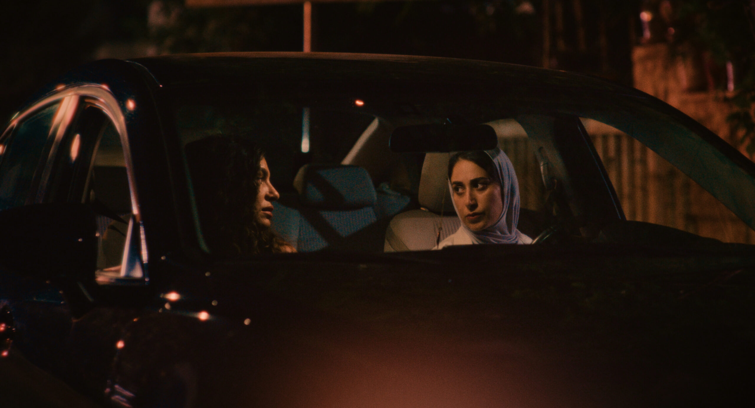 Two brown women lock eyes in the front seats of a car. One has long dark hair and the other wears a hijab. The image shows part of the car but is focused in on the front window. The lighting is dark on the outside of the car.