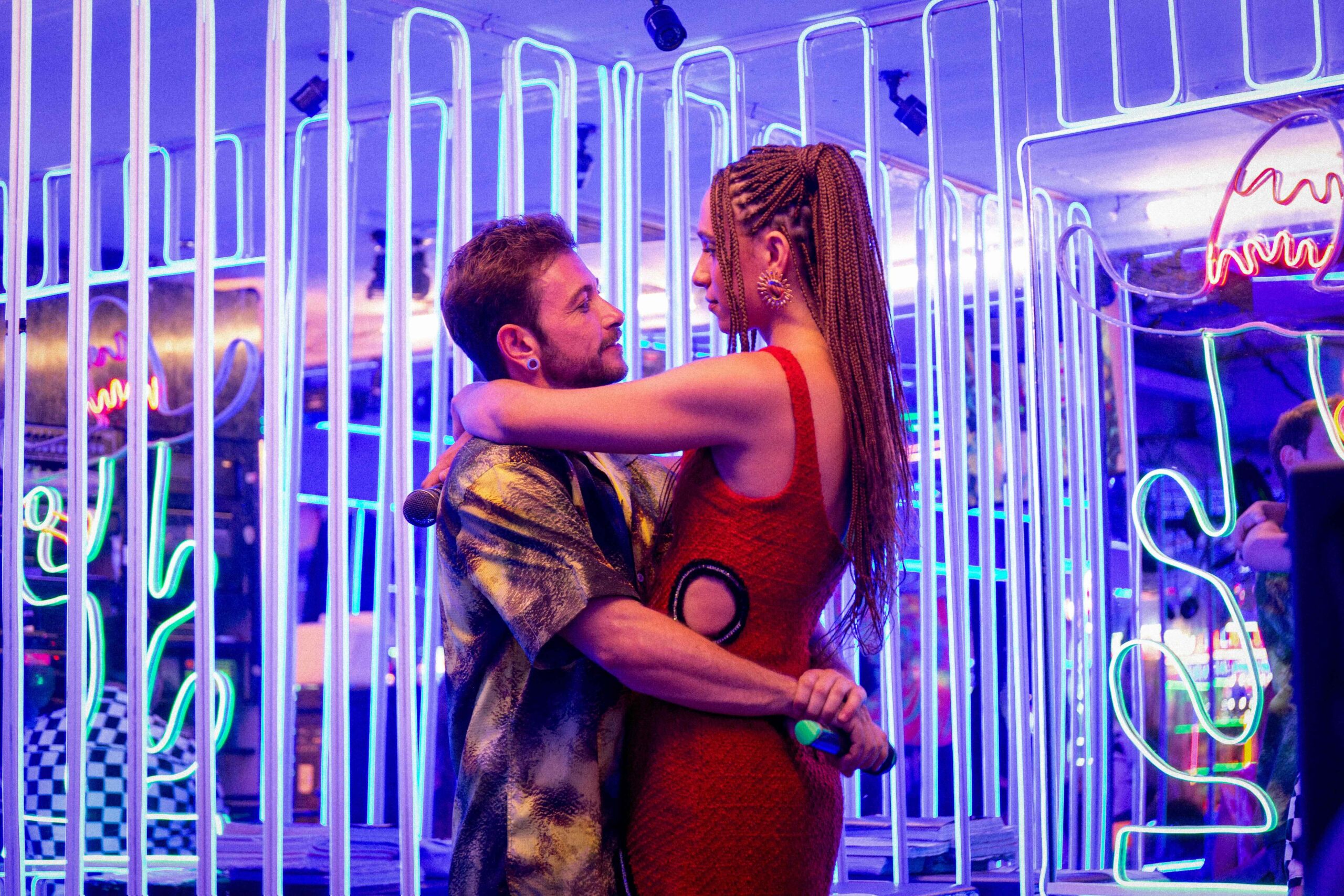 A white trans man and black trans woman embrace against a background of neon lights and iridescent metallic shapes.
