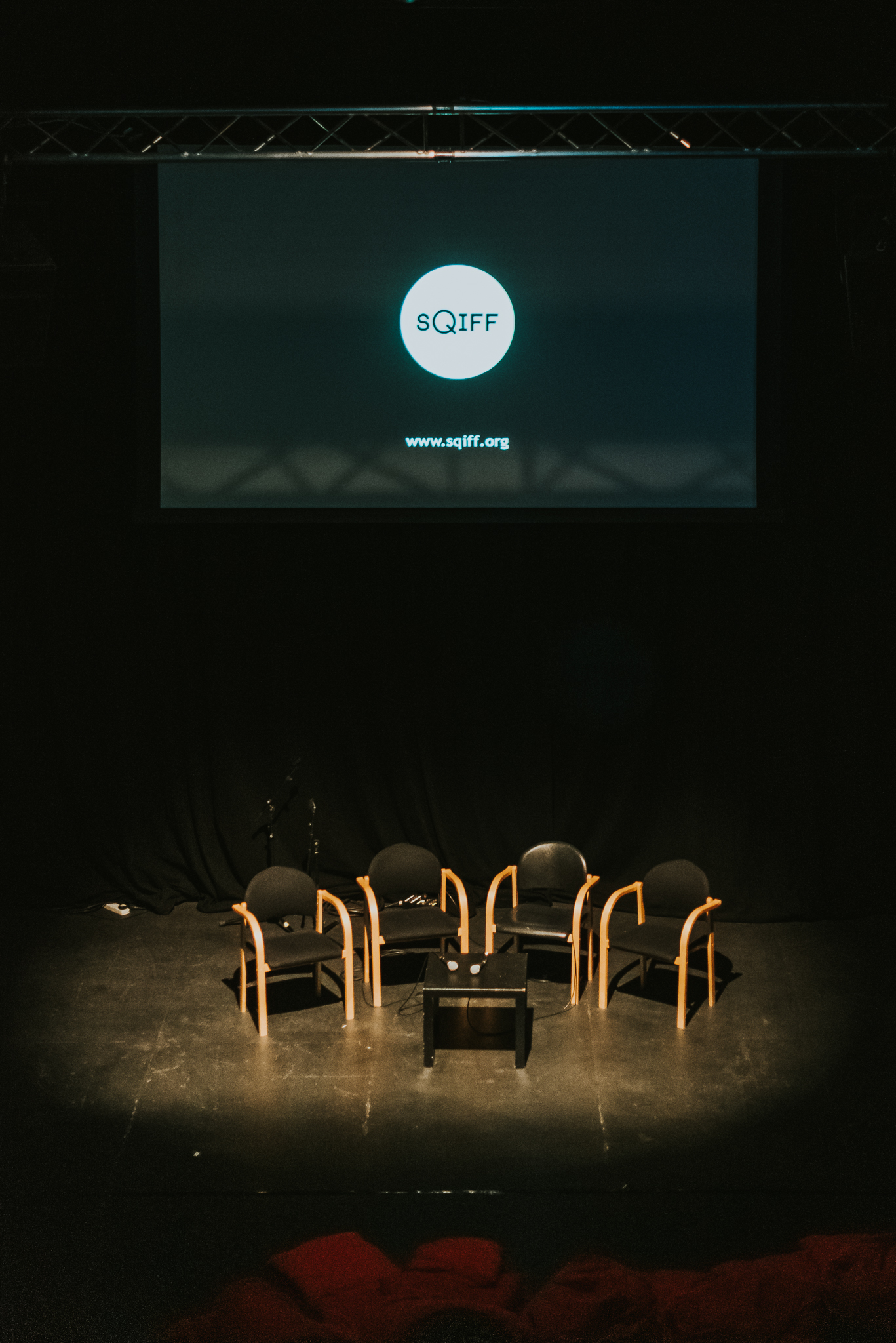 A dark room with a screen presenting the SQIFF logo. Below the screen are four empty black chairs and a coffee table with two microphones on.