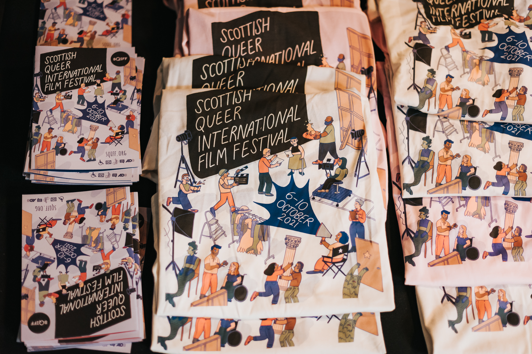 A busy table with SQIFF merch from 2021. Pictured is a stack of beige and pink tshirts with lots of characters running around a film set and the text 'Scottish Queer International Film Festival, 6-10 October 2021', and a stack of brochures with the same image and text.