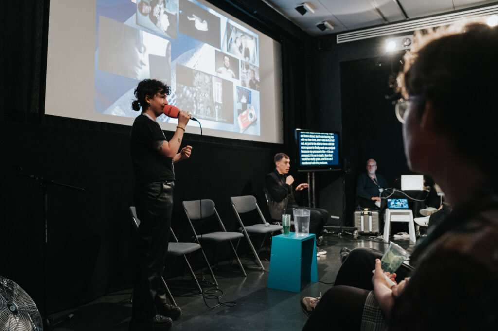 A white transmasculine person speaks into an orange microphone. There is a projection screen with a collage on behind them. There are two access workers to the right of them, one signing BSL and the other is live captioning