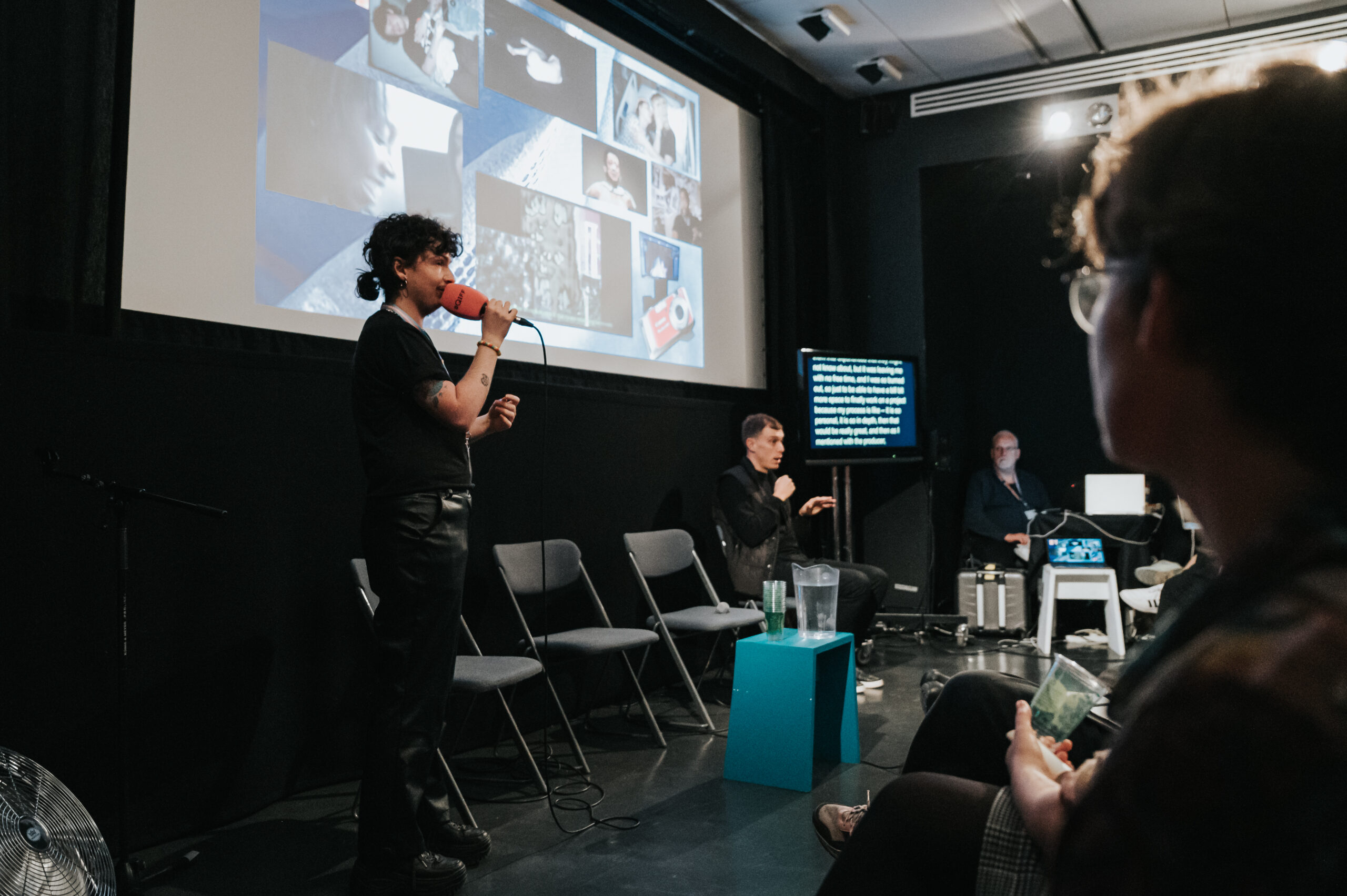 A white transmasculine person speaks into an orange microphone. There is a projection screen with a collage on behind them. There are two access workers to the right of them, one signing BSL and the other is live captioning