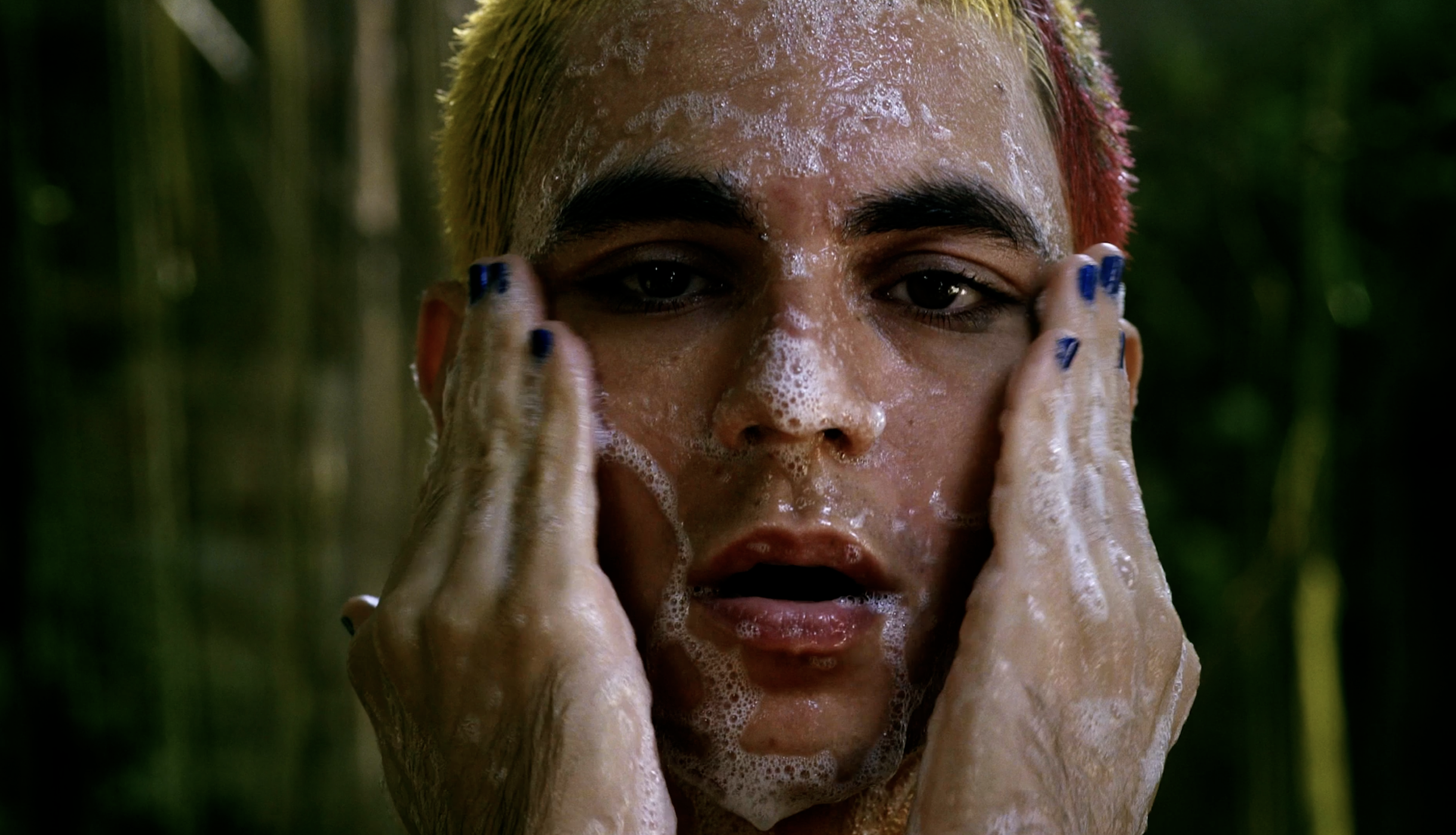The character (Renê) with blue-painted fingernails washes their soapy face in a tight shot, gazing forward with parted lips while hands frame their cheeks in front of soft, out-of-focus foliage.