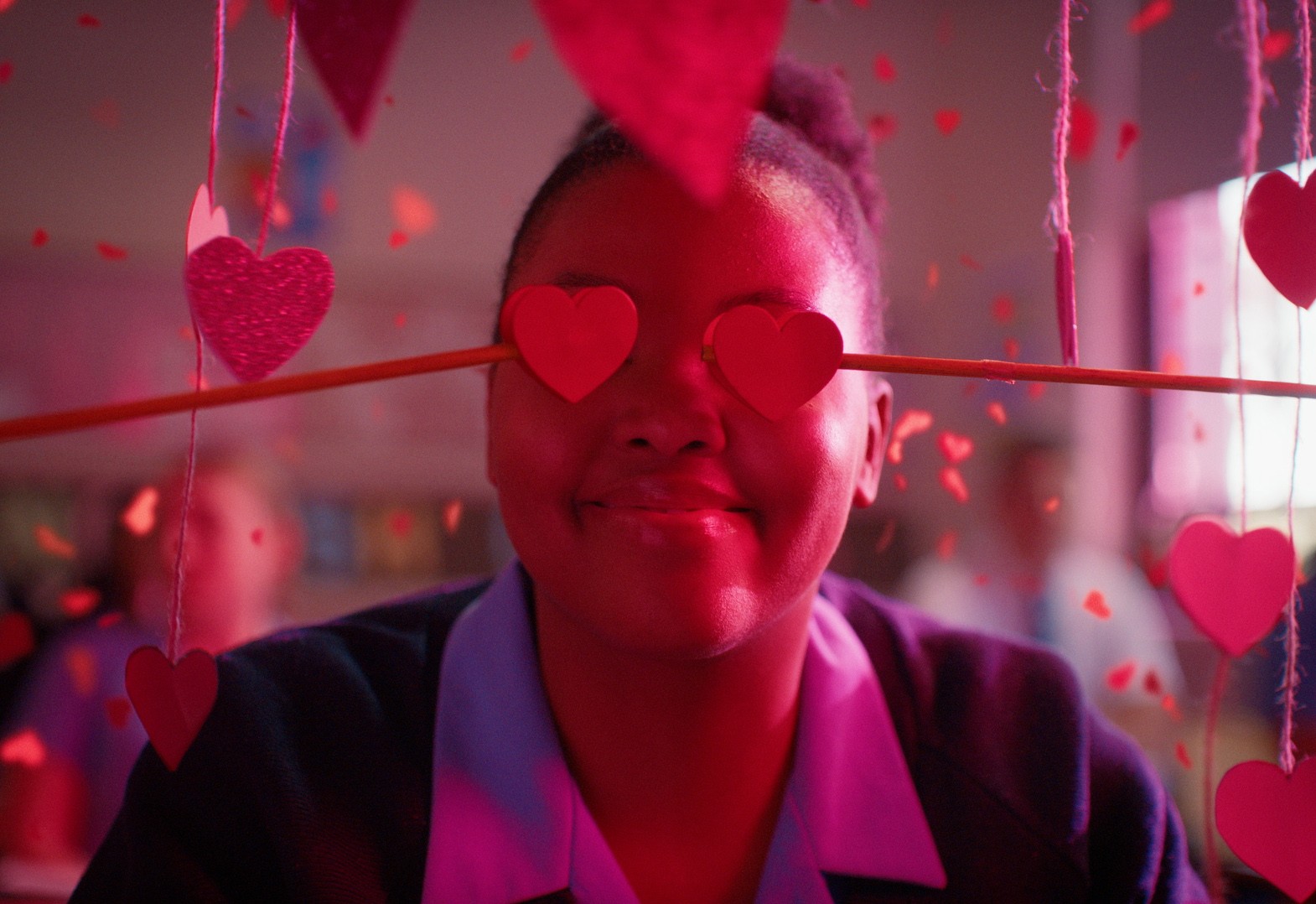 A pink close-up of a Black girl smiling facing the camera with paper crafted heart shaped glasses and hearts all over the image.